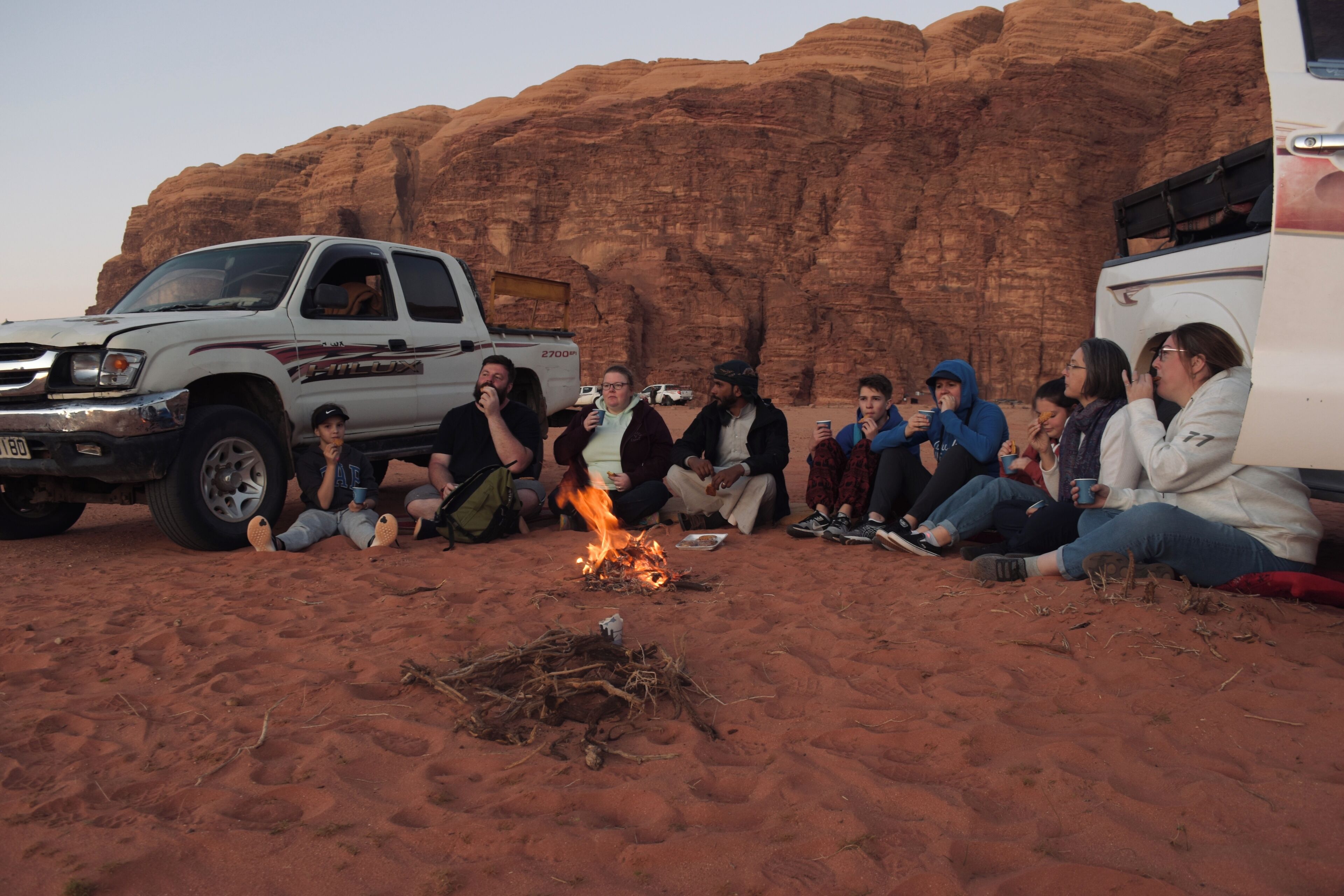 A group of people sitting around a campfire in a desert setting with rocky cliffs in the background.