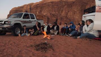 A group of people sitting around a campfire in a desert setting with rocky cliffs in the background.