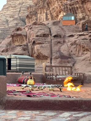 A desert scene with a fire, a wooden bench, a patterned rug, and a small building in the background.