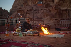 A desert scene with a person sitting by a fire, surrounded by traditional furnishings and a hookah. - Magic Bedouin Star - Campsite (Wadi Rum)