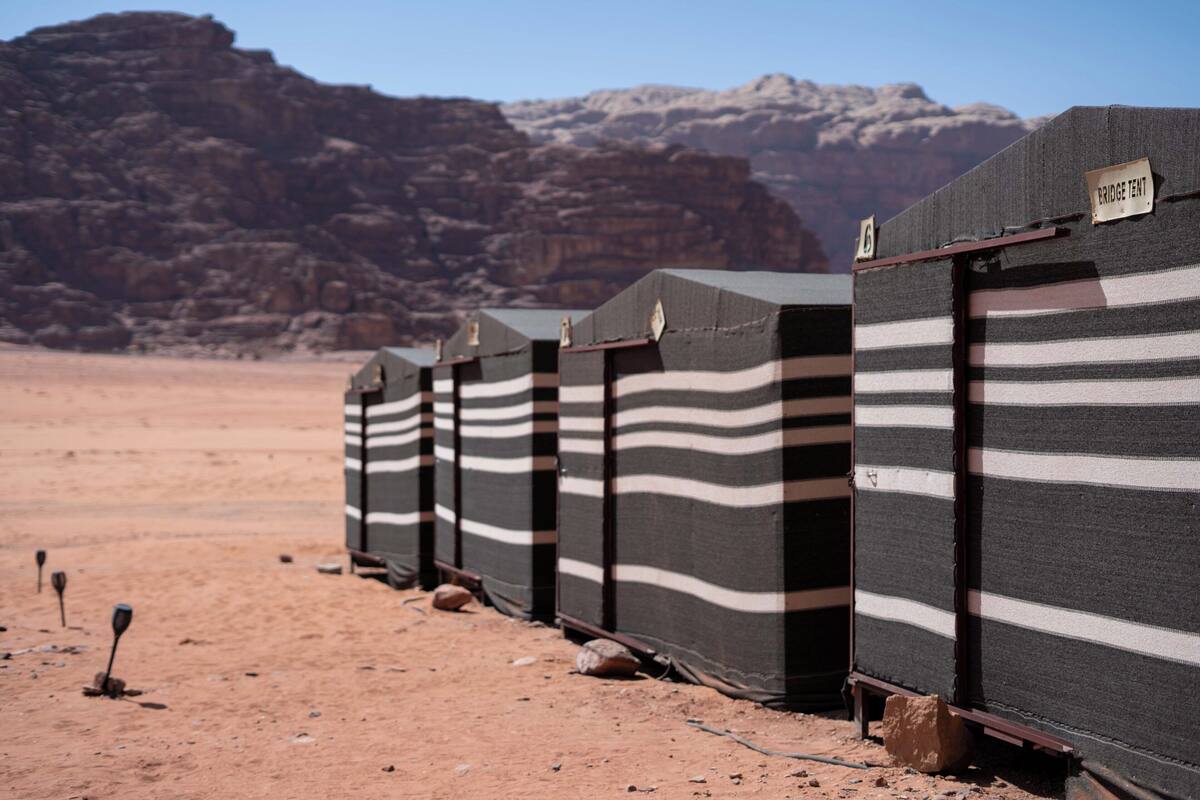 Una hilera de tiendas a rayas con la etiqueta 'BRIDGE TENT' en un paisaje desértico con montañas rocosas al fondo.