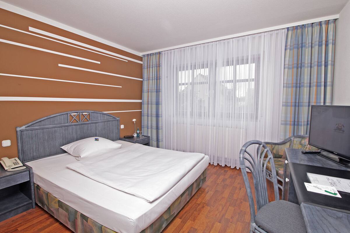 A hotel room with a bed, a desk with a computer, a chair, a telephone, and a window with curtains.