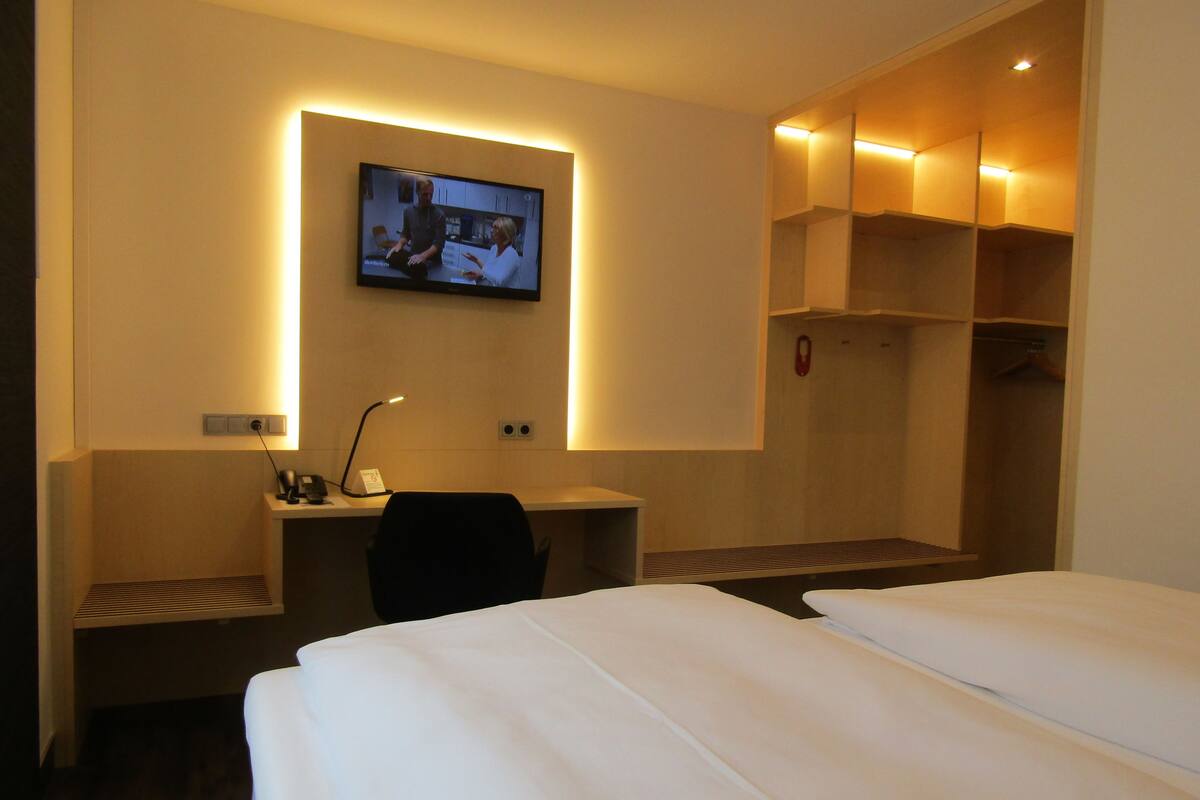 A hotel room with a bed, a desk with a chair, a TV mounted on the wall, and a wardrobe.