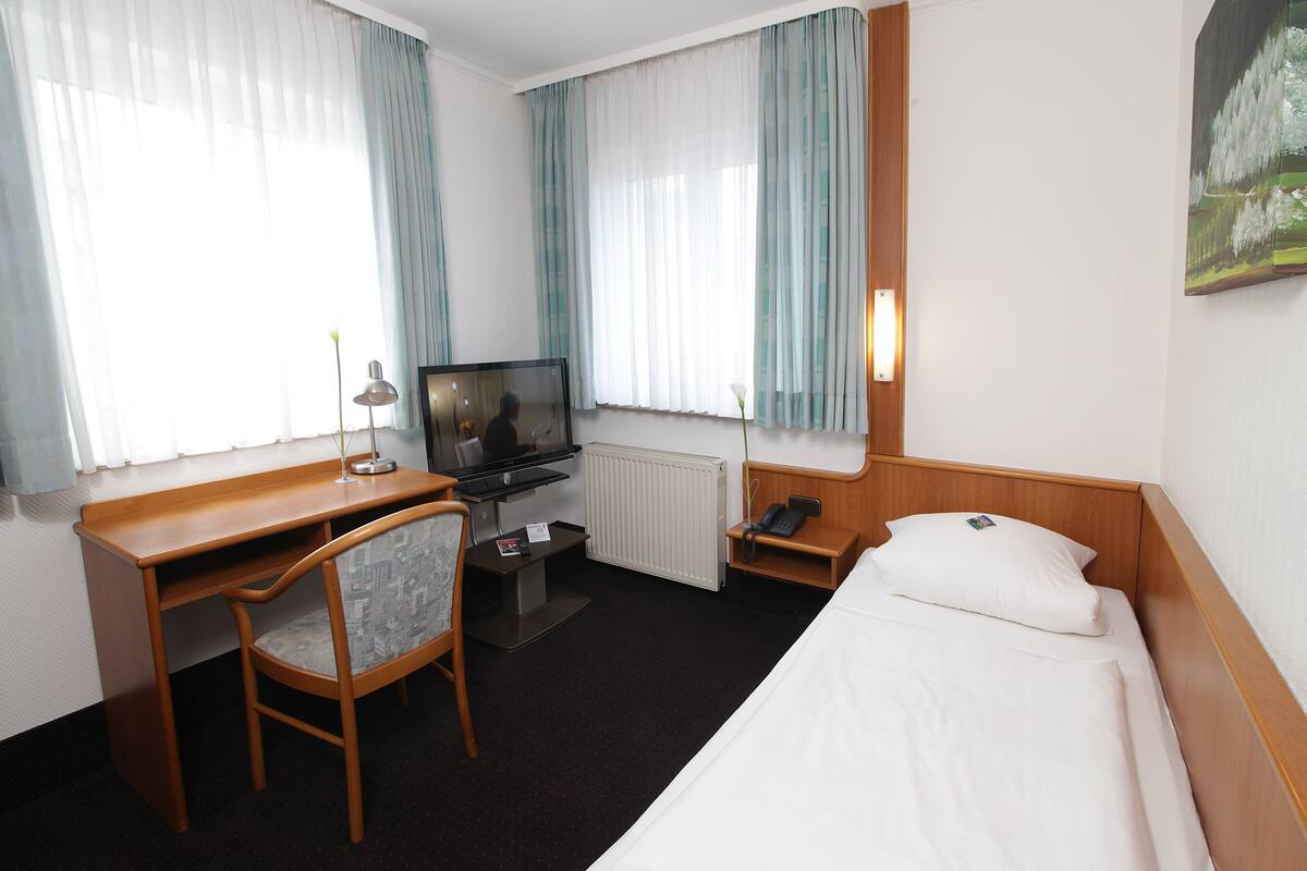A hotel room with a wooden desk, a chair, a bed, a TV, and a painting on the wall.