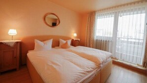 Una habitación de hotel con una cama grande, dos mesitas de noche con lámparas, un espejo redondo en la pared y una ventana con cortinas traslúcidas.