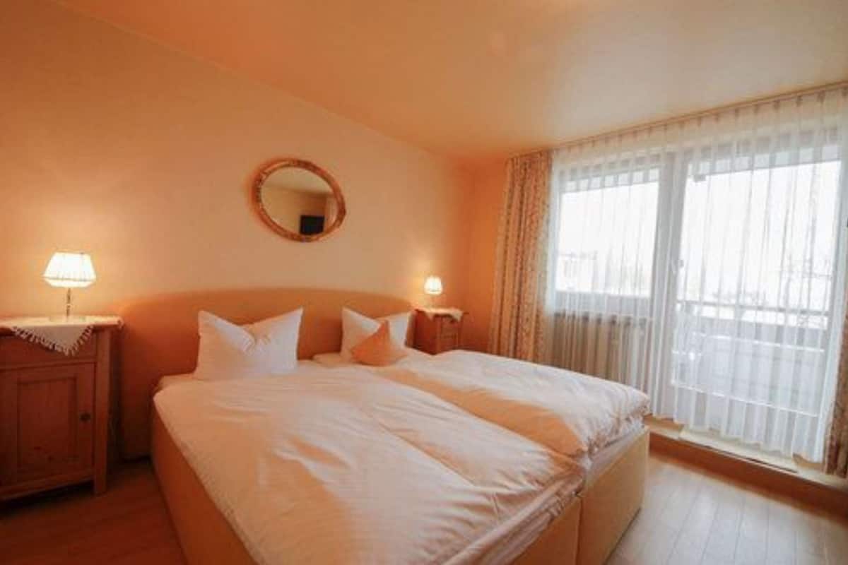 Una habitación de hotel con una cama grande, dos mesitas de noche con lámparas, un espejo redondo en la pared y una ventana con cortinas traslúcidas.