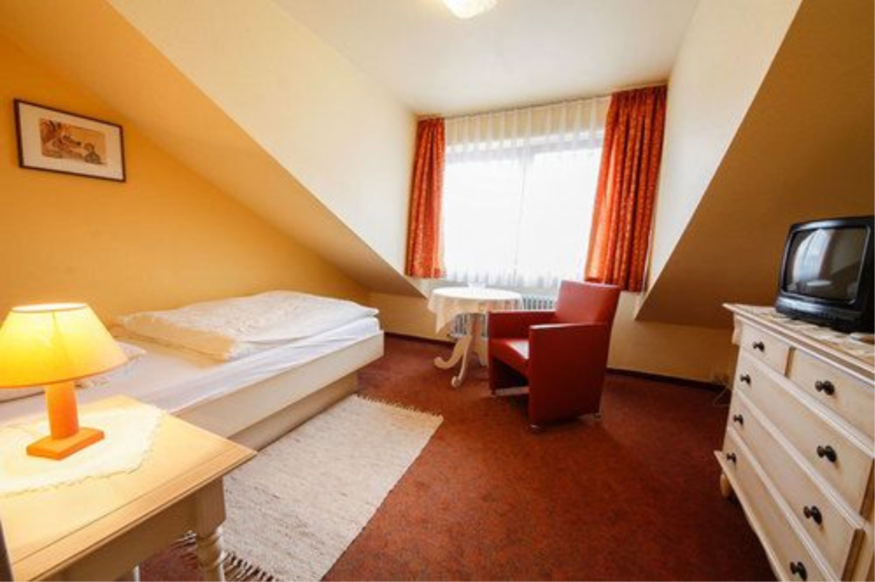 A small, triangular room with a bed, a desk with a lamp, a chair, a dresser, and a television.