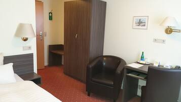 A hotel room with a bed, a desk, a chair, a wardrobe, a lamp, and a framed picture on the wall.