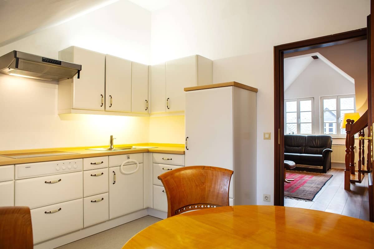 A compact kitchen with white cabinets, a wooden dining table, and a view into a living room with a black sofa and a staircase.