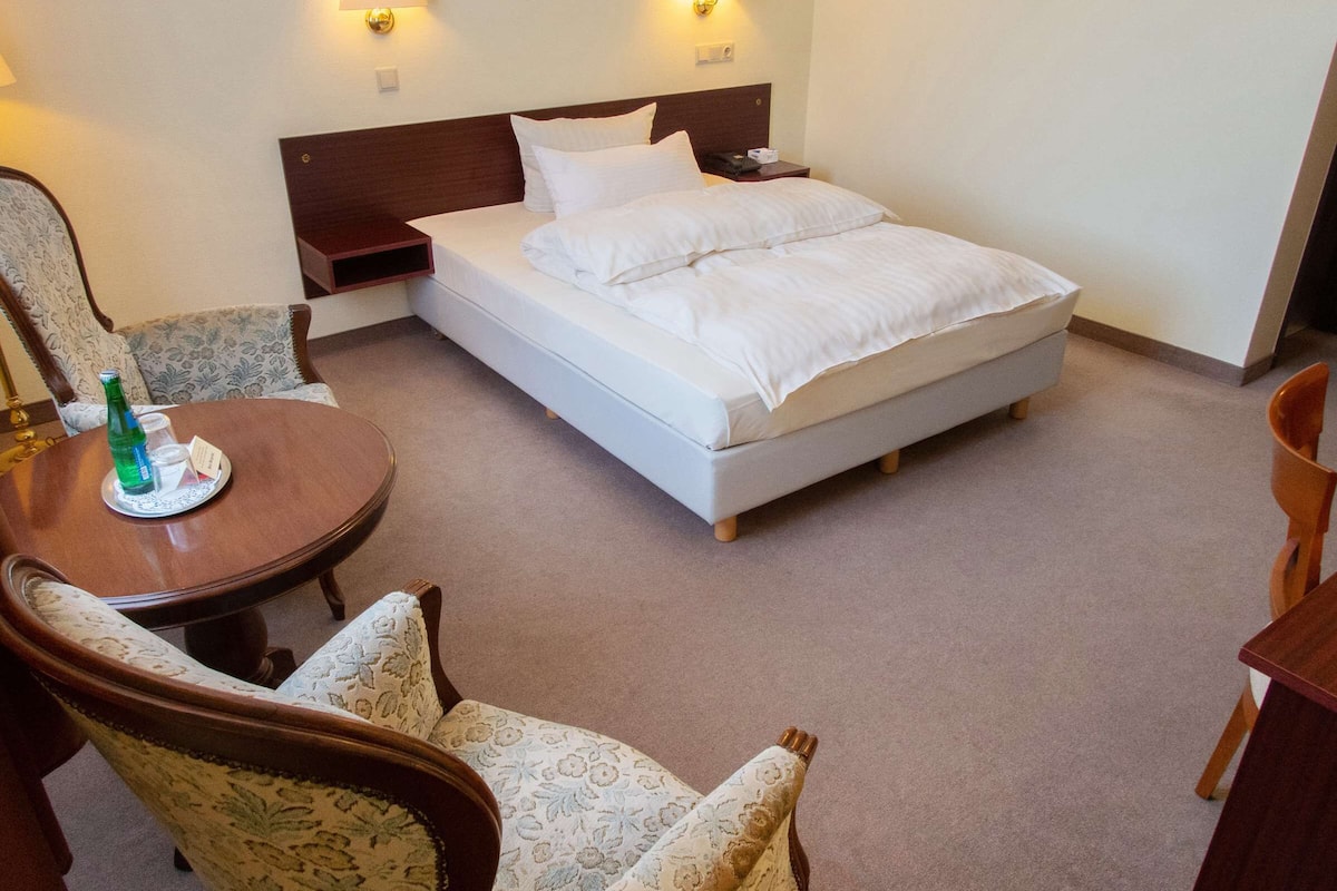 A hotel room with a single bed, two bedside tables, a small table with a bottle and glasses, and two patterned armchairs.
