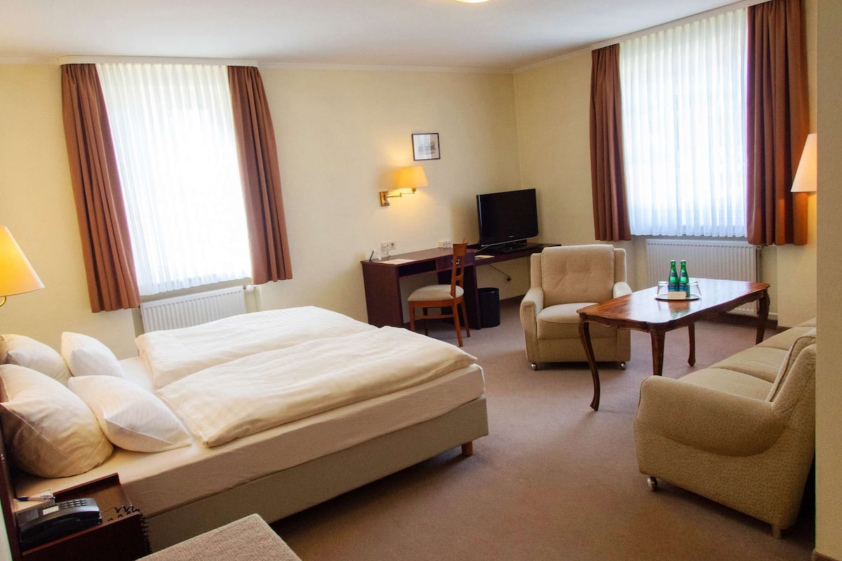 A hotel room with a large bed, a desk with a computer, a chair, a small table with bottles, and a sofa.