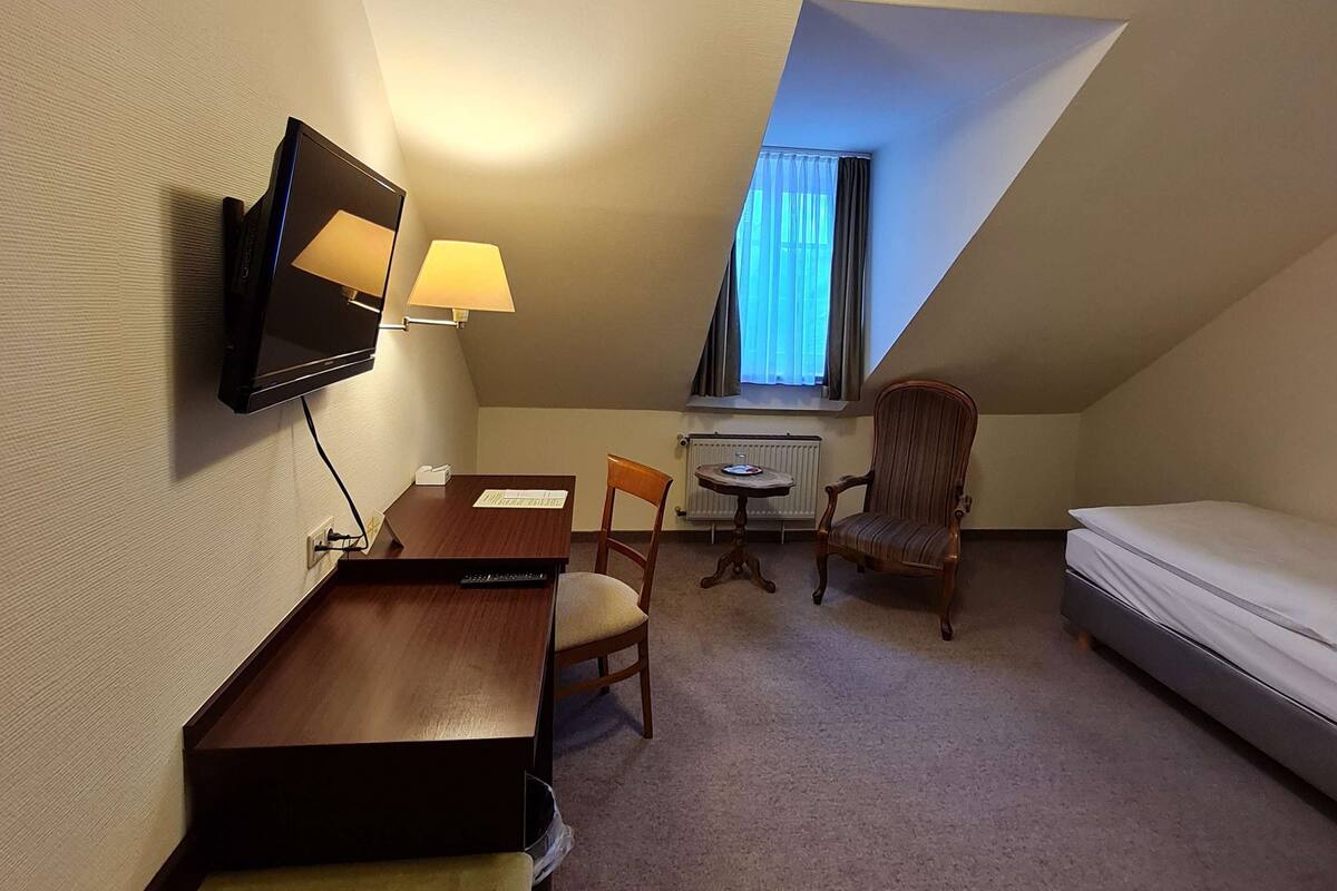 A small, triangular room with a bed, a desk with a lamp, a chair, and a television mounted on the wall.