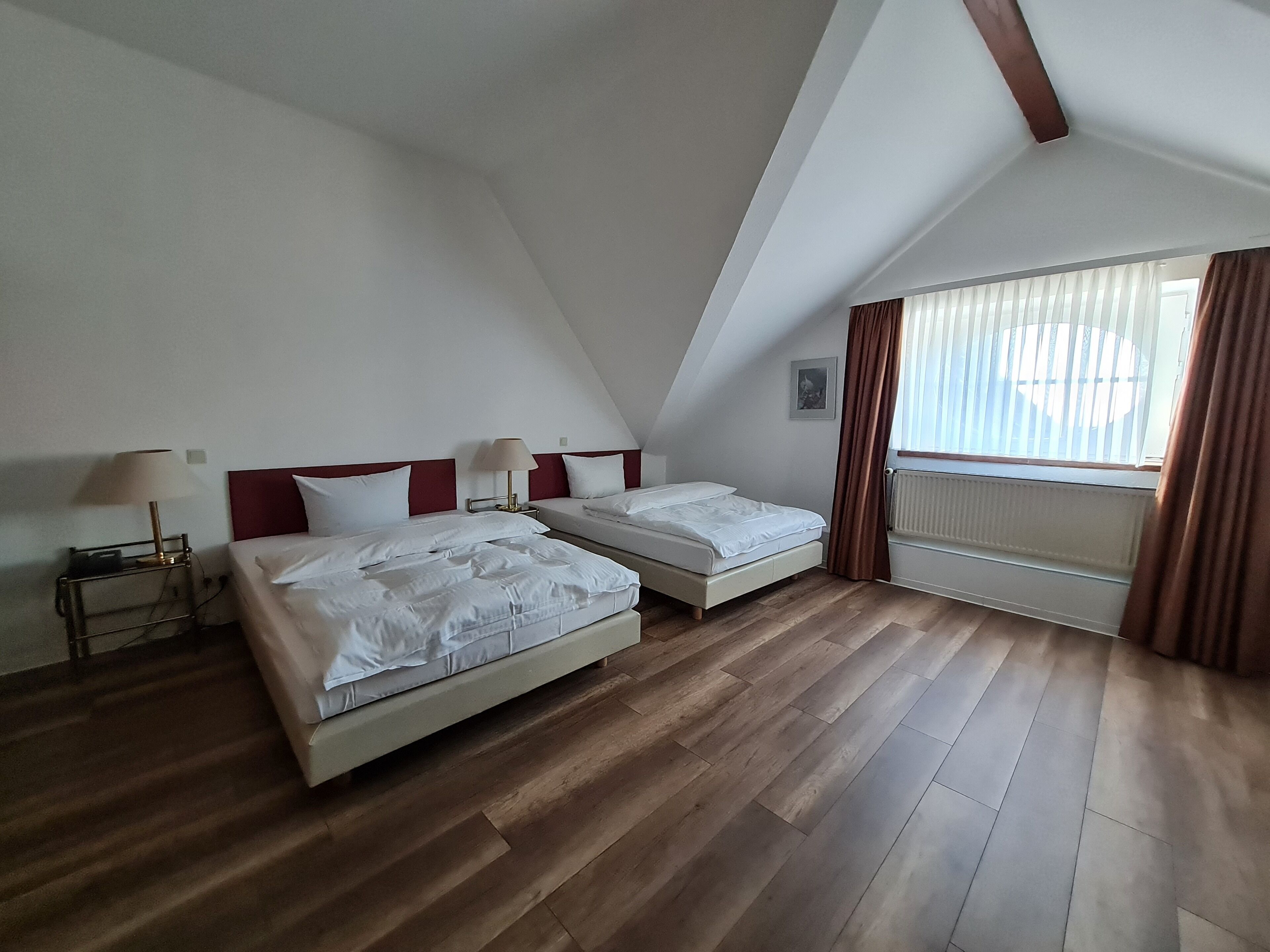 A room with two beds, a radiator, a window with curtains, and a lamp on a bedside table.