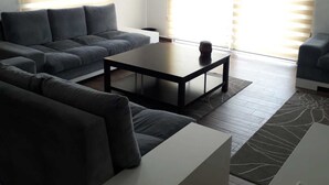 Un salon moderne avec un canapé d’angle, une table basse et une petite table d’appoint blanche. Il y a deux fenêtres équipées de stores, un luminaire suspendu et un tapis à motifs sur le sol.