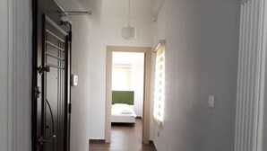 Un couloir étroit avec un plancher en bois, des murs blancs et une porte sur la gauche. On aperçoit au loin un lit avec une tête de lit verte.