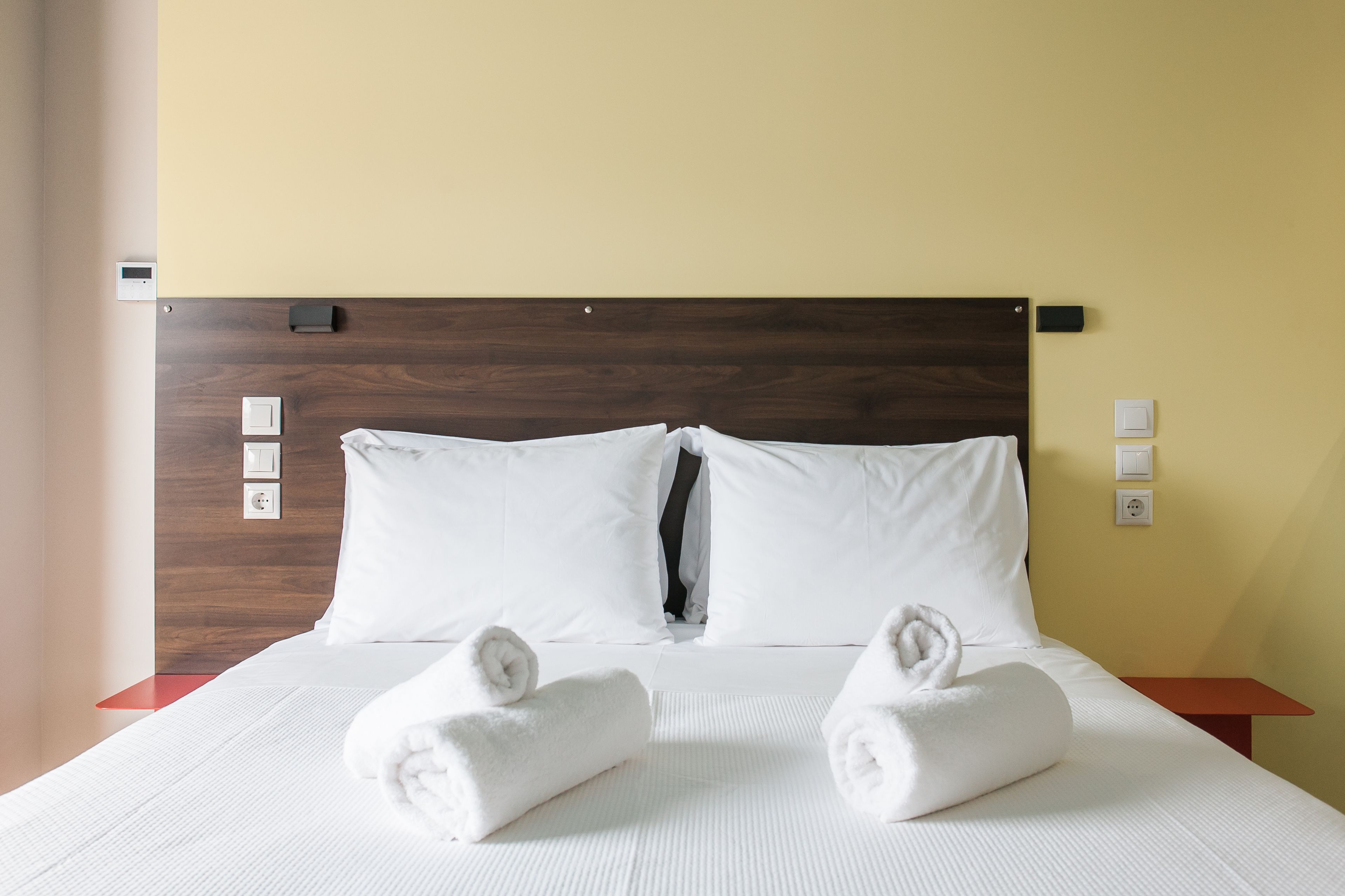 A neatly made bed with white linens and two rolled towels, a wooden headboard, and a small bedside table with a red surface.