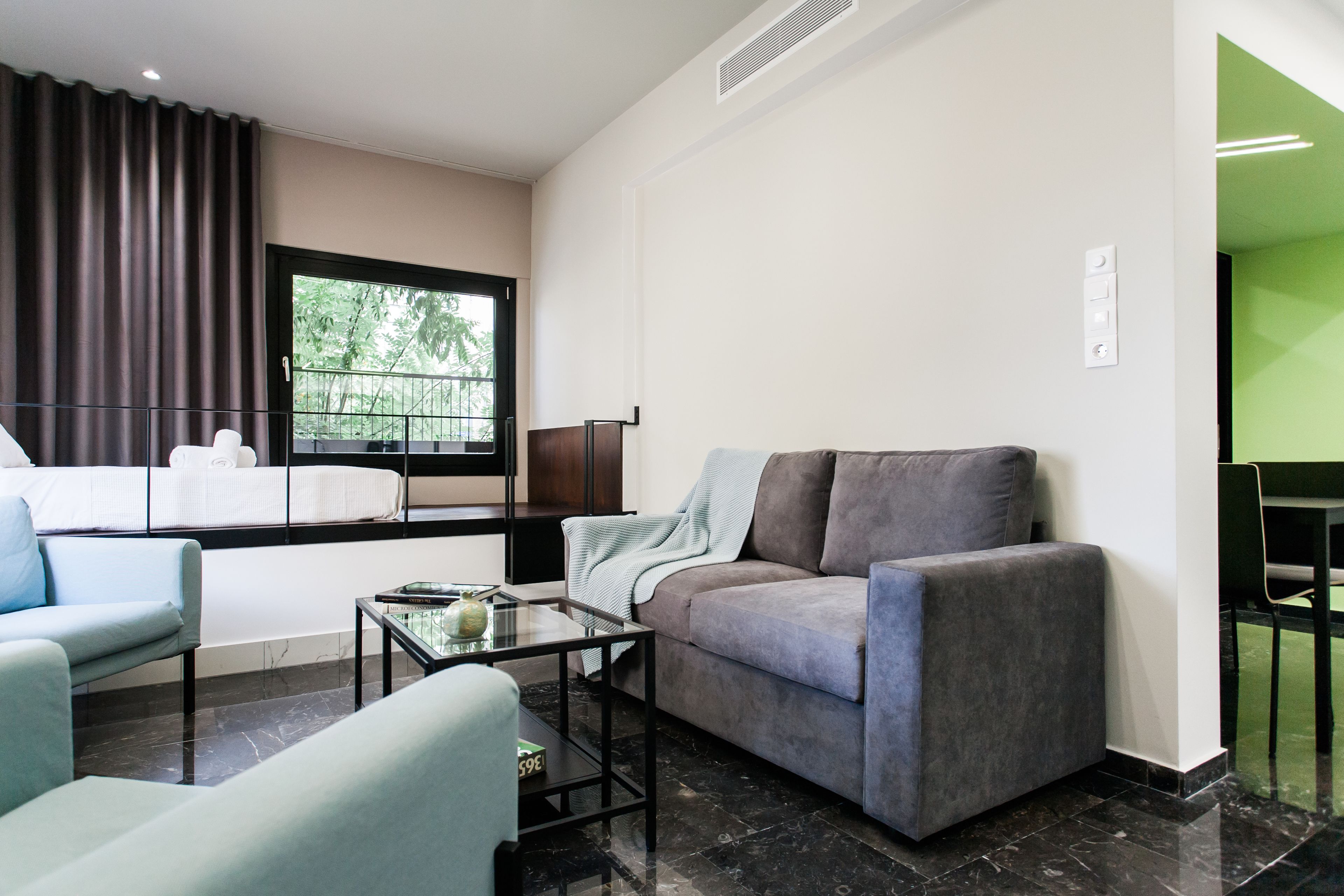 A modern living room with a grey sofa, a blue armchair, and a glass coffee table. There is a window with curtains, a black floor, and a green wall visible in the background.