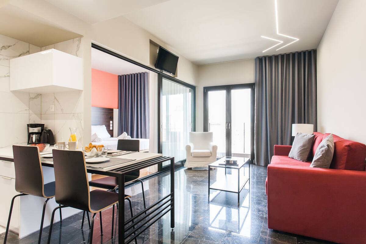 Una sala de estar moderna con un sofá rojo, un sillón blanco y una mesa de centro de vidrio. Hay un comedor con una mesa y sillas negras, y una cocina con gabinetes blancos y un salpicadero de mármol.