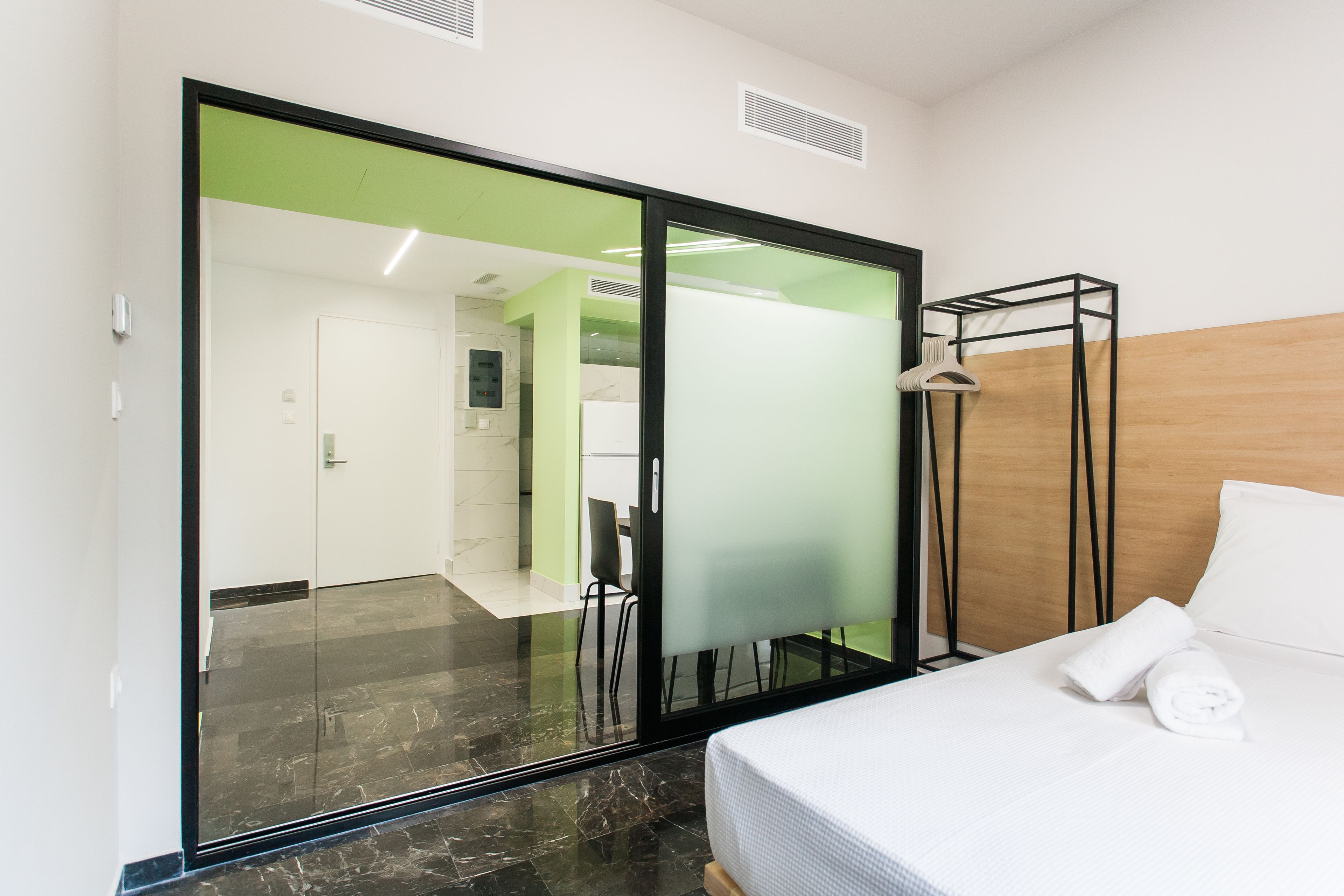 A modern hotel room with a large sliding glass door leading to a bathroom, a neatly made bed with a white comforter, and a wooden headboard.