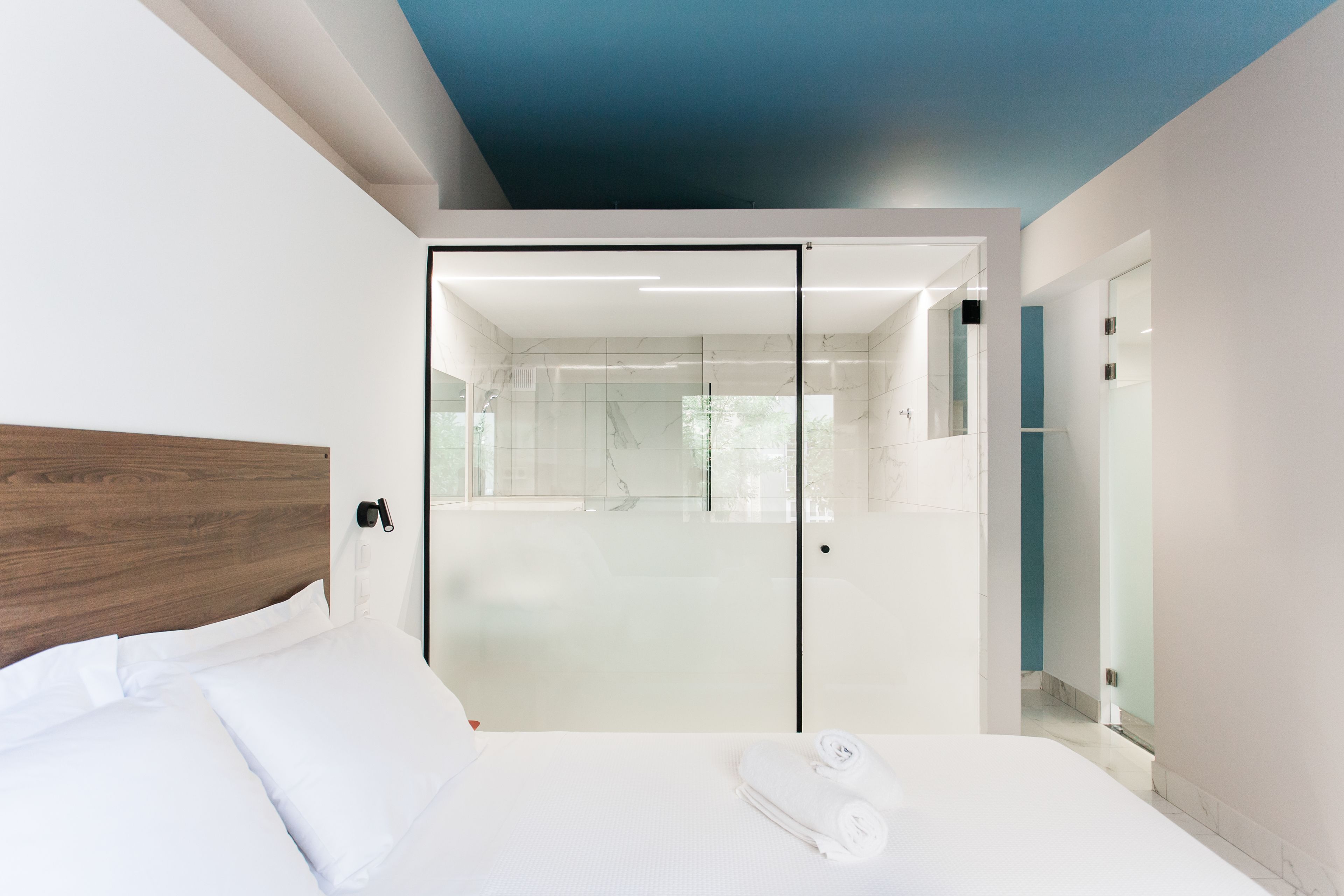 A modern hotel room with a wooden headboard, a large bed with white linens, a glass shower enclosure, and a view of the adjacent bathroom.