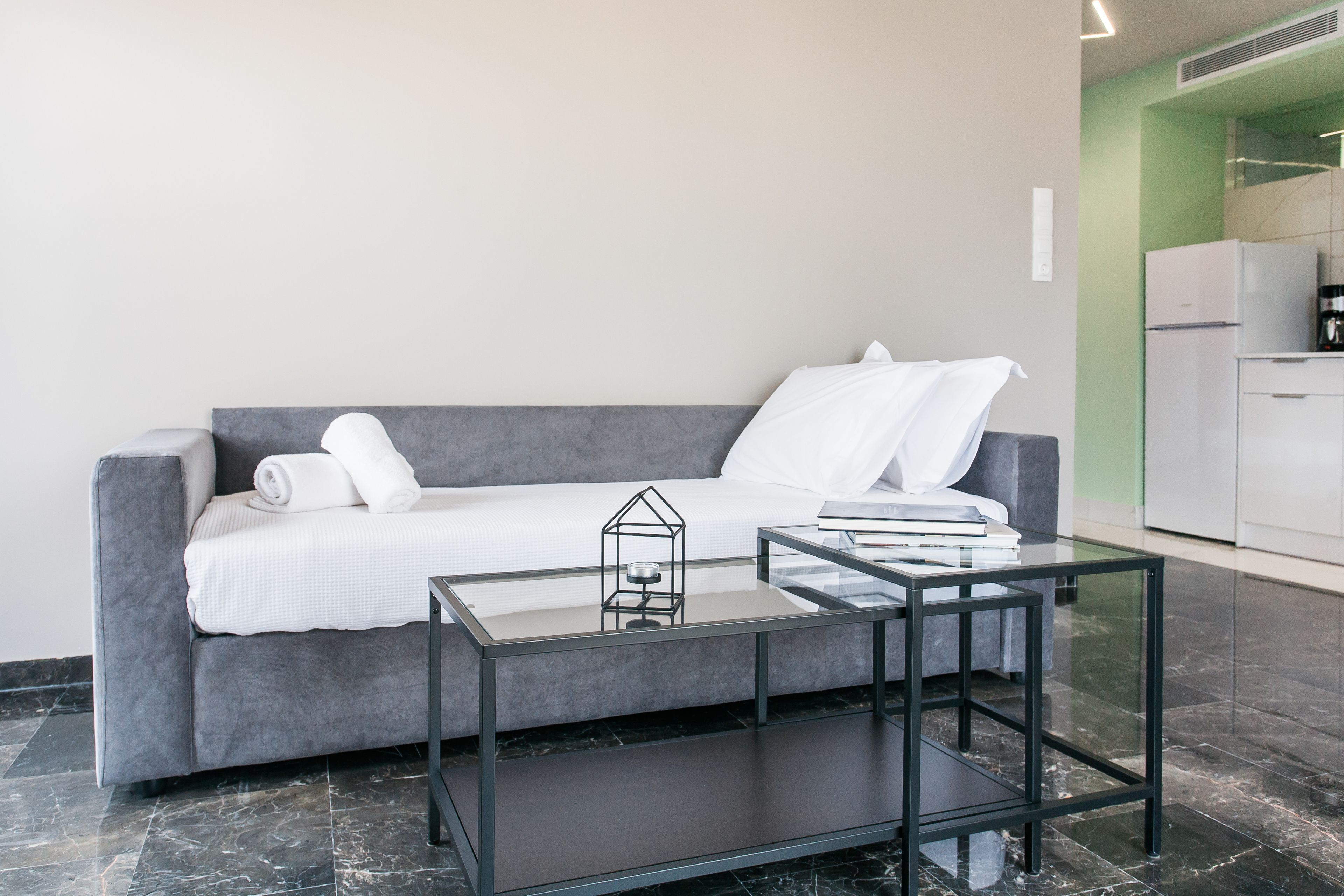 Een moderne woonkamer met een grijze bank, een glazen salontafel, en een keuken in de achtergrond.