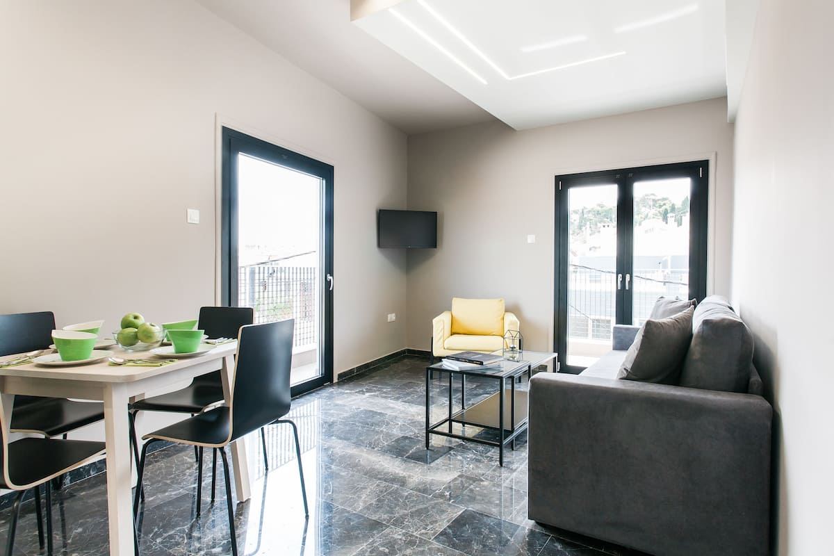 Una sala moderna con un sofá, una mesa de comedor con sillas, un televisor de pantalla plana en la pared y un balcón con barandilla.