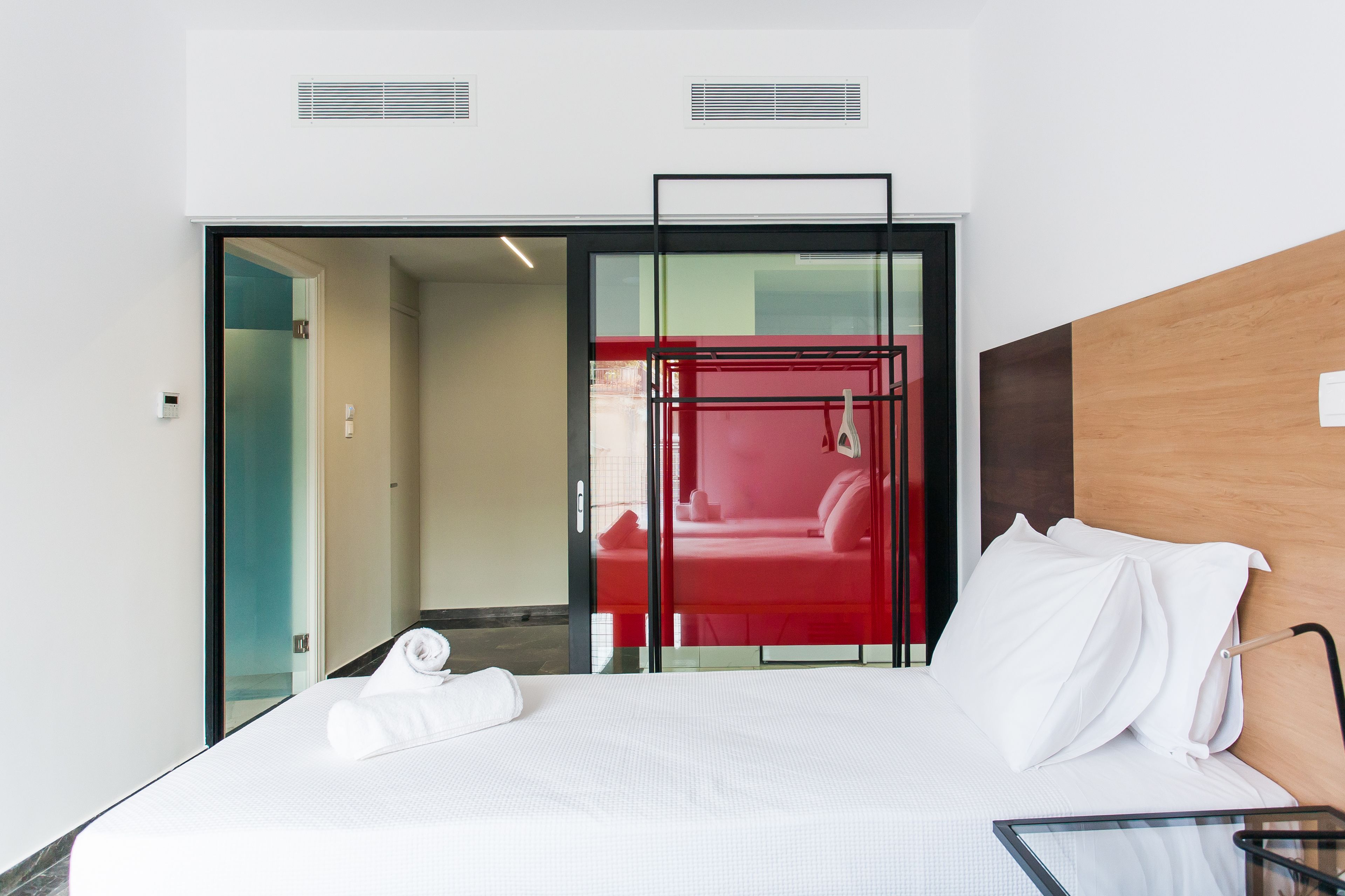 A modern hotel room with a large bed, a glass table, and a red room in the background.