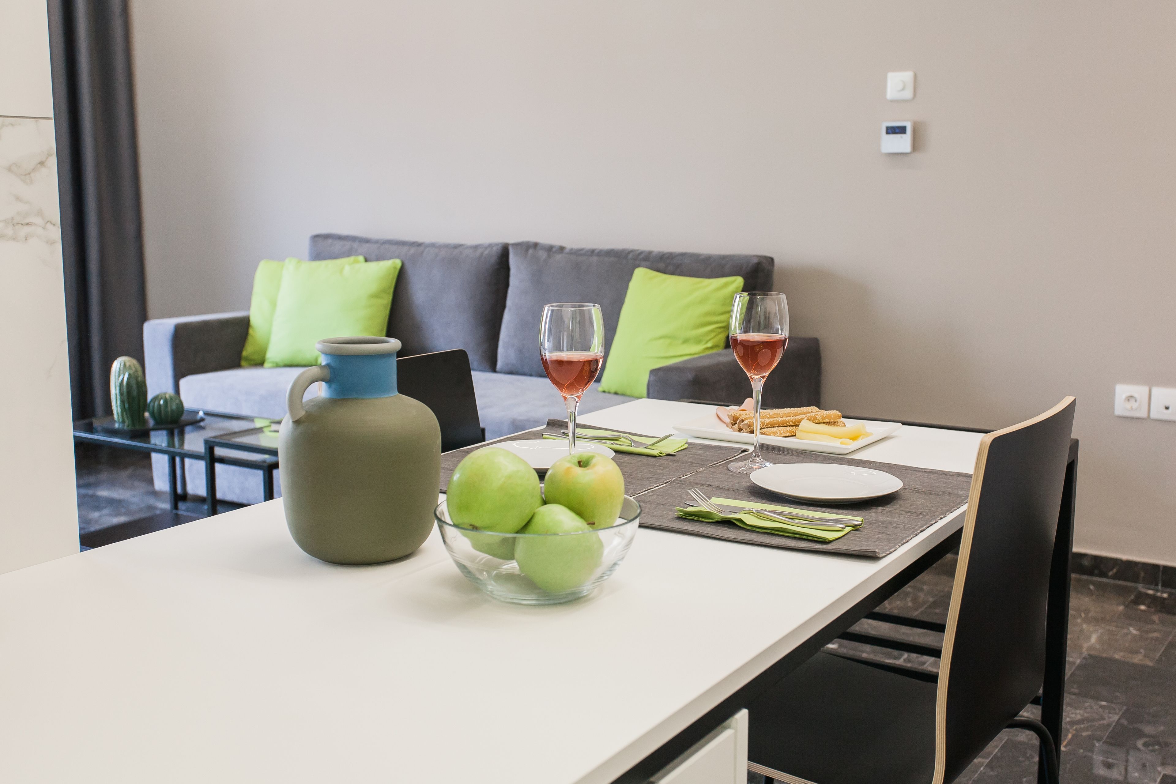 A dining table with a bowl of green apples, two wine glasses, and a blue pitcher.