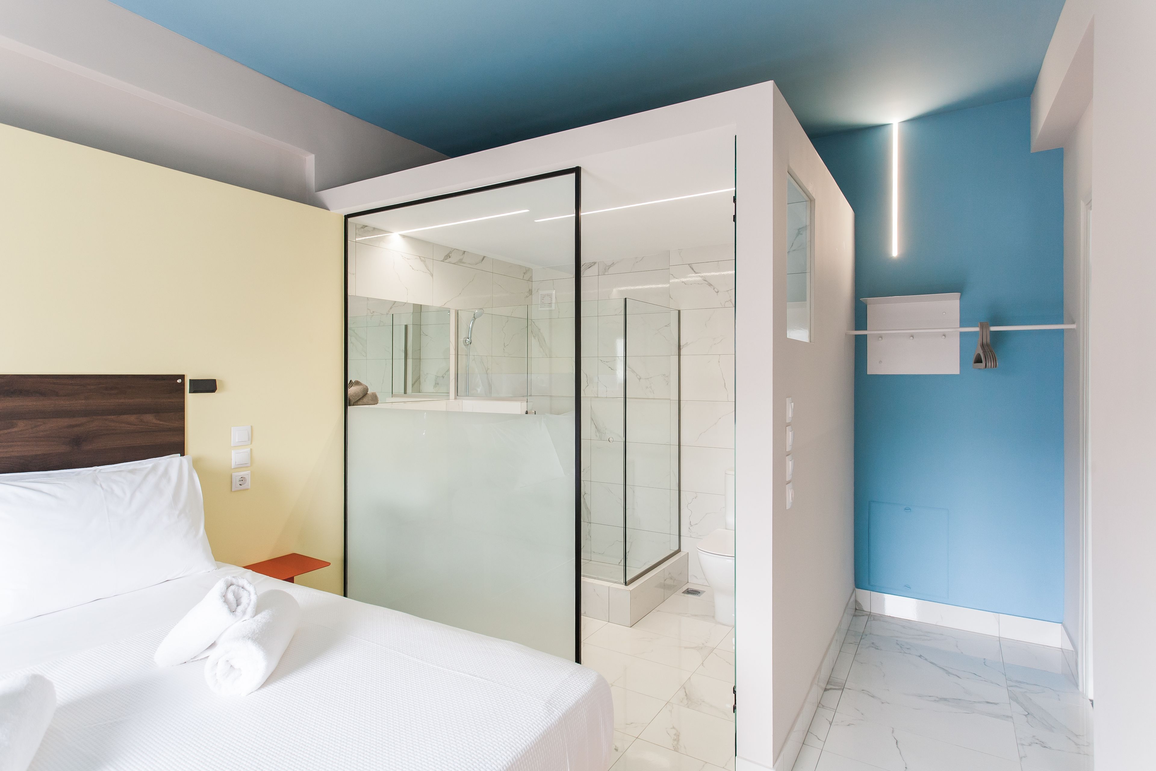 A modern hotel room with a bed, a glass-enclosed shower, a white bathroom, and a blue accent wall.