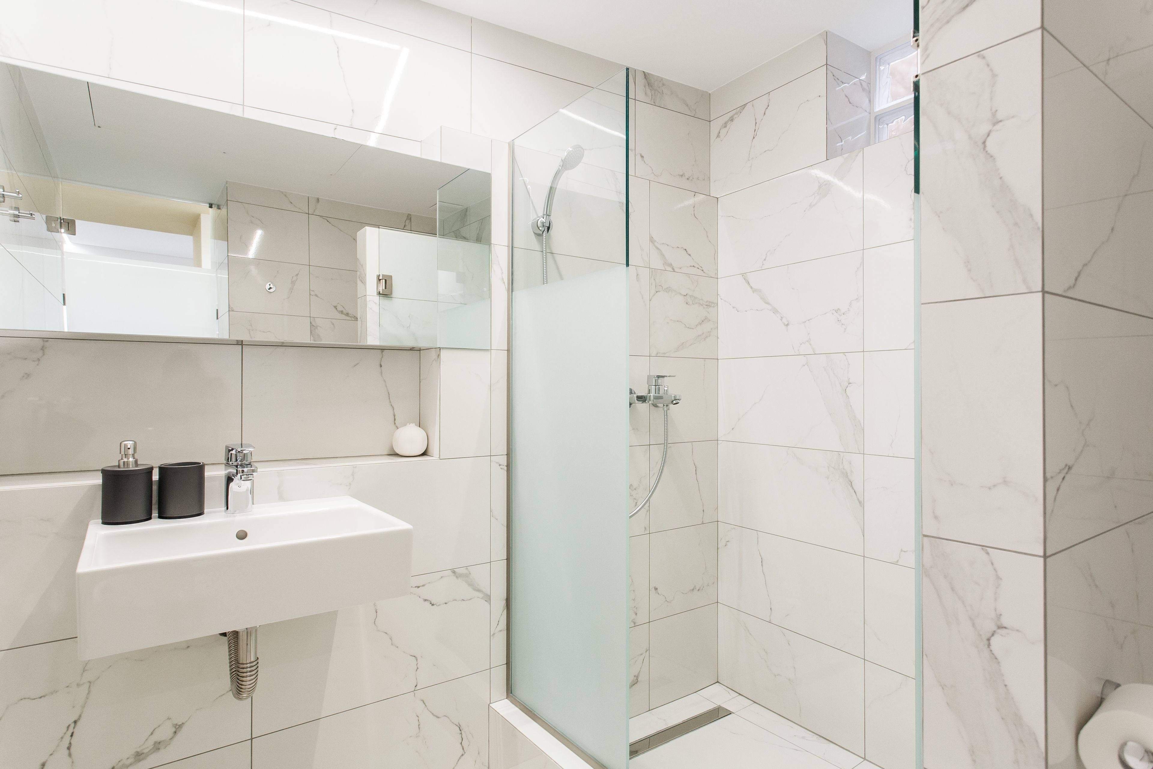 A modern bathroom with a glass-enclosed shower, a white sink with two black soap dispensers, and a marble-tiled wall.