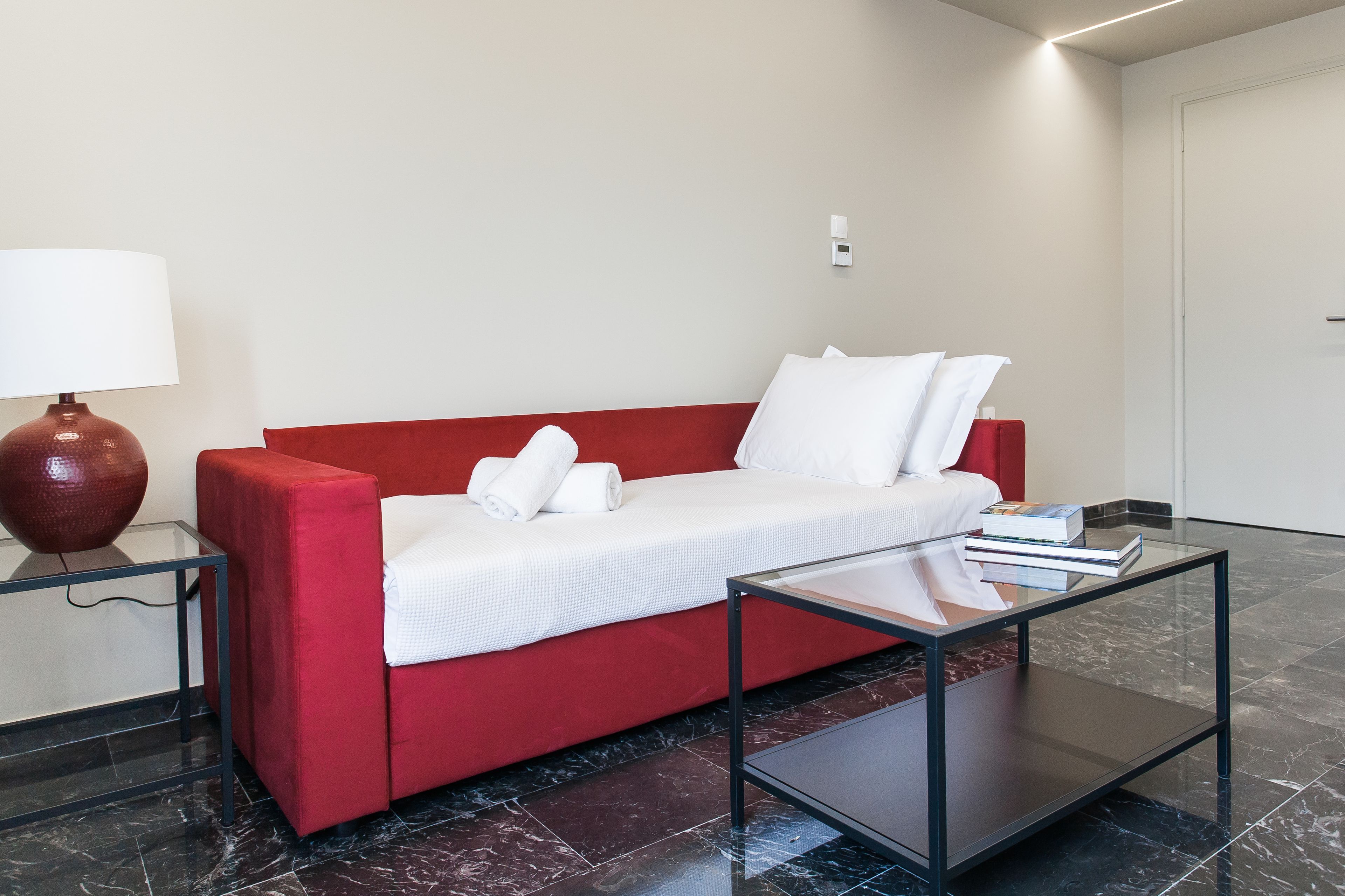 A red sofa with white bedding, a glass coffee table, and a lamp on a side table.