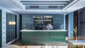A modern bar area with a green bar counter, shelves of bottles, and a seating area with wicker chairs and a table.