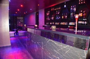Una zona de bar con mostrador de mármol, estantes llenos de botellas y un vaso decorativo.