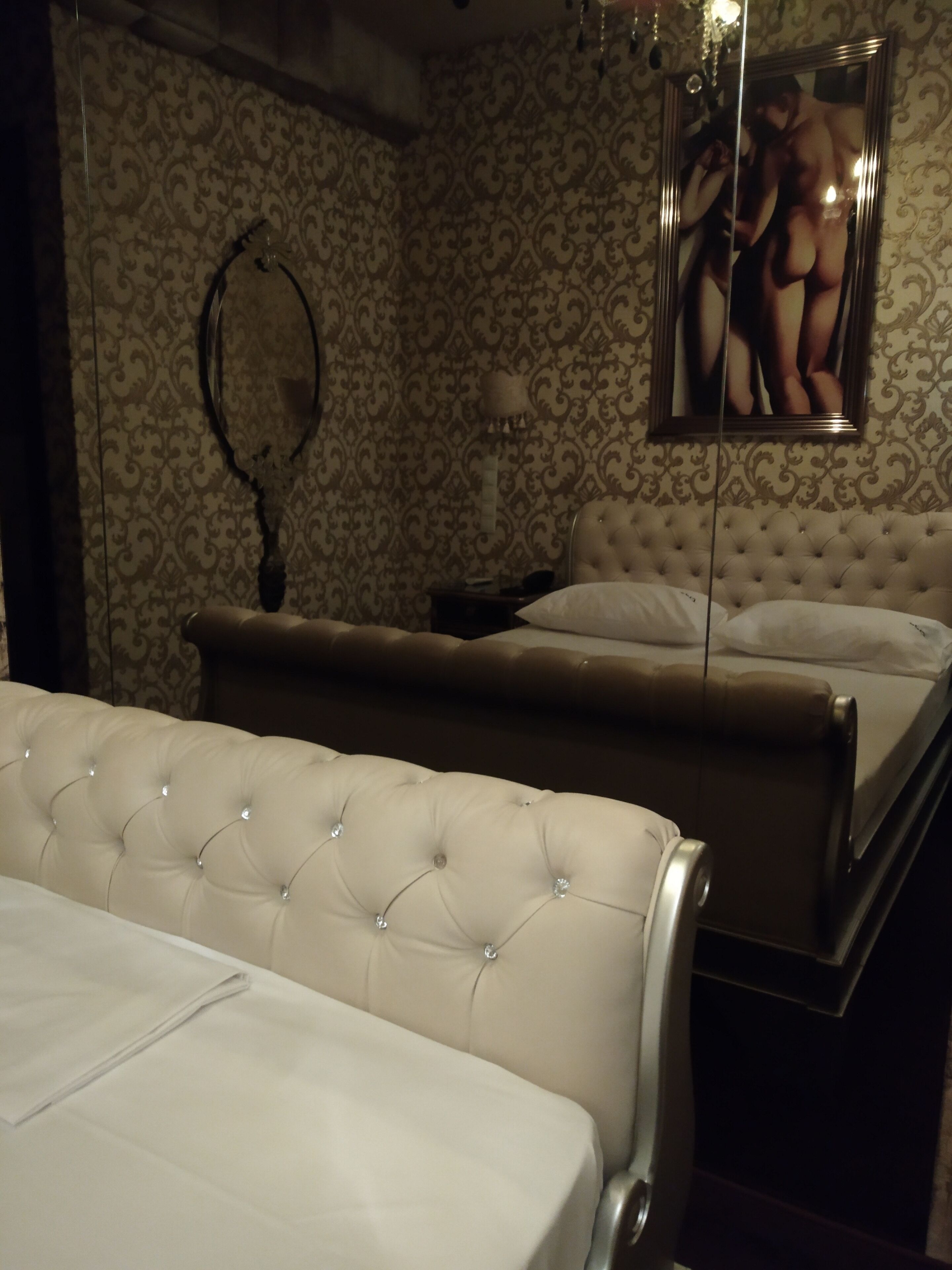 Une chambre à coucher avec une tête de lit capitonnée, un lit recouvert de draps blancs, une table de chevet et un miroir dans lequel se reflète une personne.