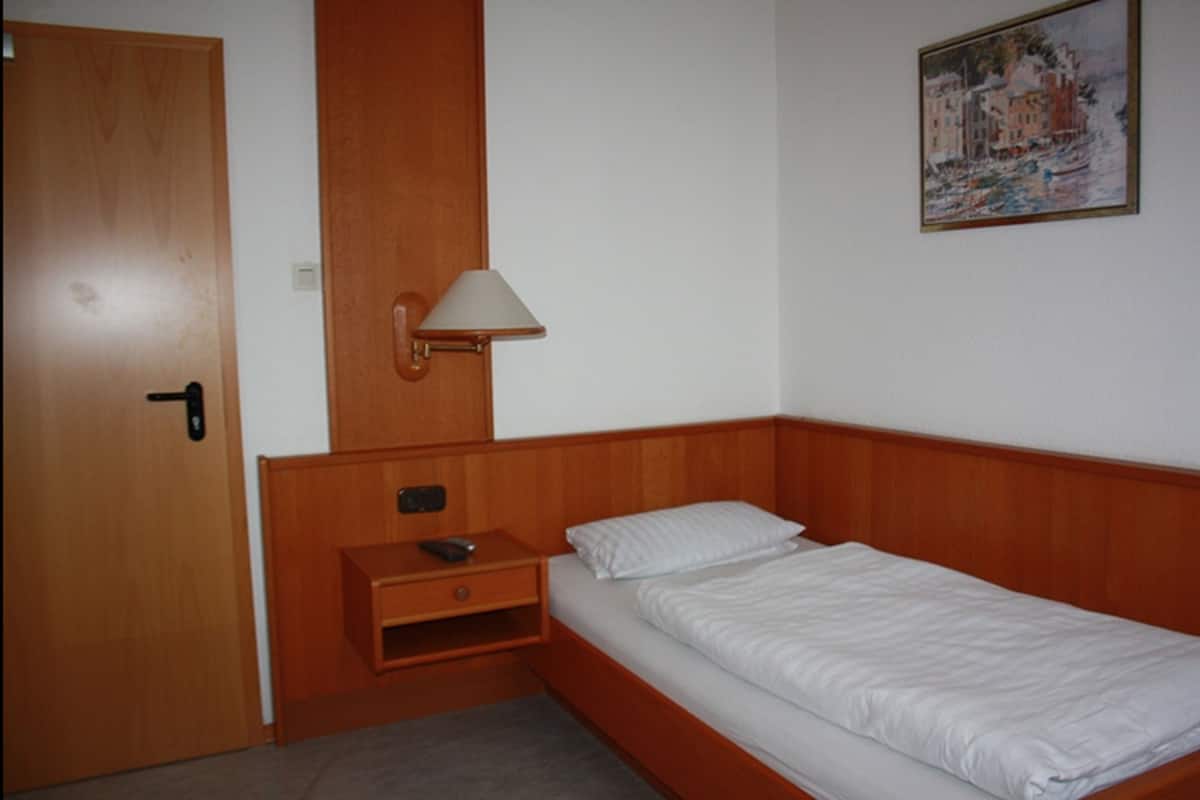 A single bed with a wooden headboard, a bedside table with a lamp, a small drawer, and a door.