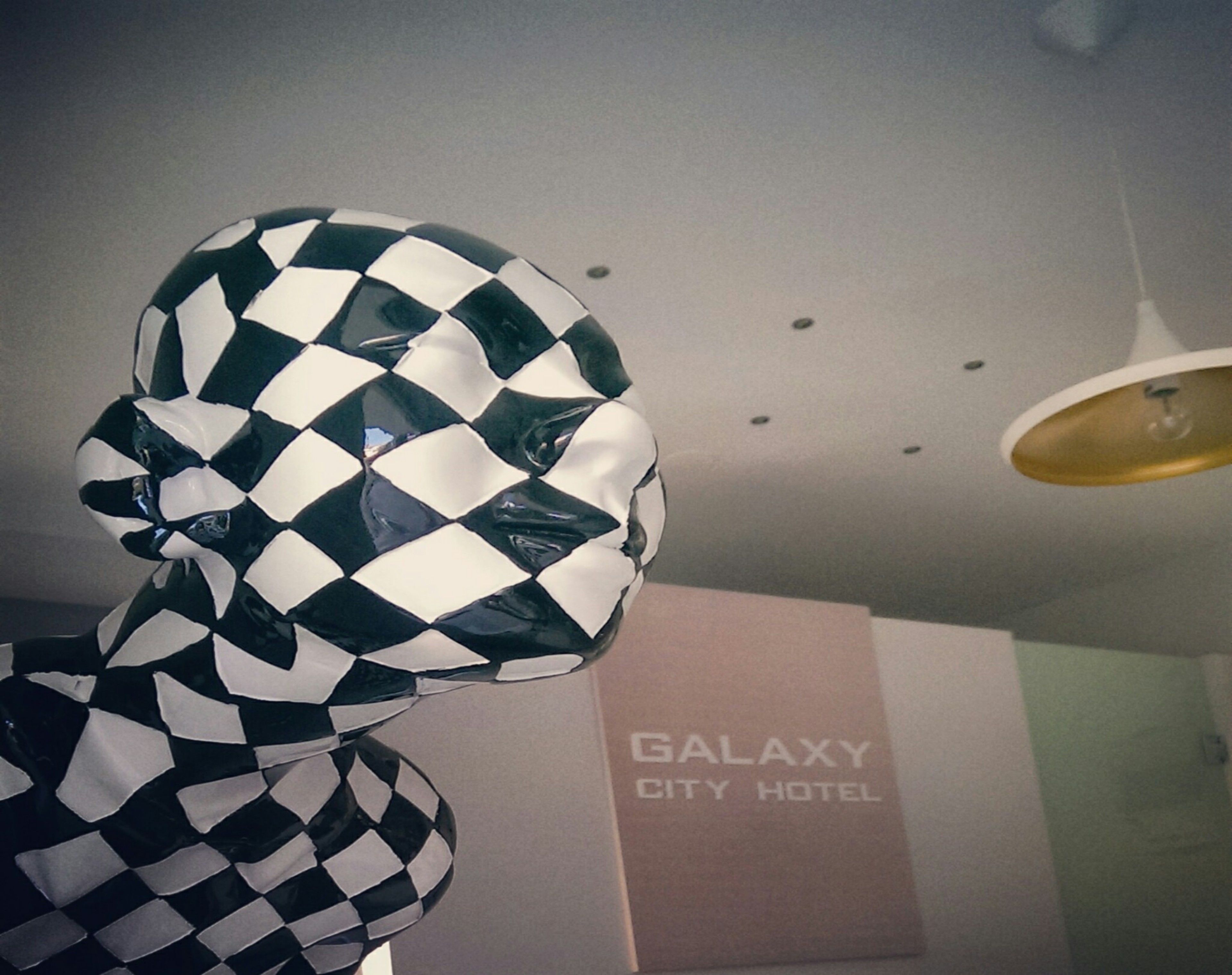 Una habitación con un letrero del Hotel Galaxy City en la pared, una escultura a cuadros y una lámpara en el techo.
