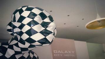 Una habitación con un letrero del Hotel Galaxy City en la pared, una escultura a cuadros y una lámpara en el techo.