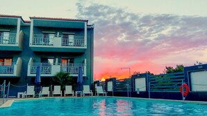 Una piscina con sillas de descanso y sombrillas, un edificio con balcones, y un atardecer de fondo.