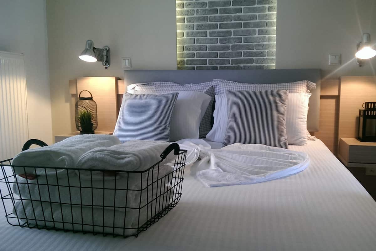 Un lit bien fait, avec du linge de lit blanc, un panier à linge en métal et un mur de briques avec une fenêtre au-dessus.