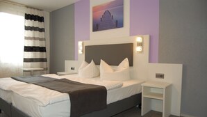 A neatly made bed with pillows and a blanket, a bedside table, a nightstand, a wall-mounted light, and a painting above the bed. - Hotel Schneider (Lich)