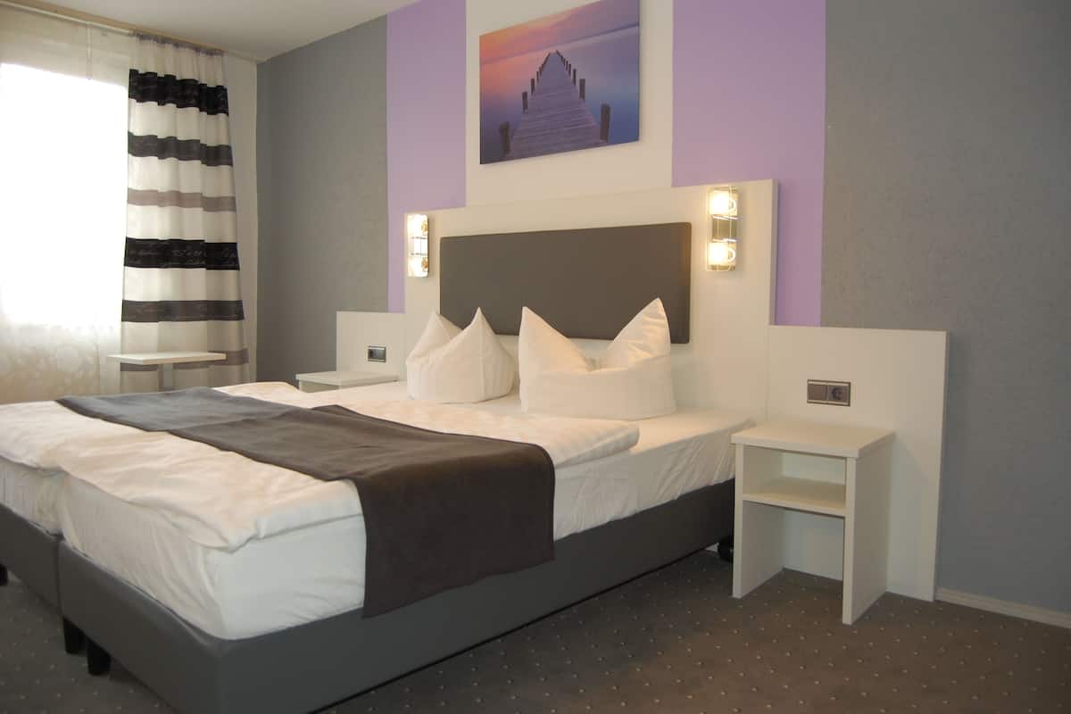 A neatly made bed with pillows and a blanket, a bedside table, a nightstand, a wall-mounted light, and a painting above the bed.