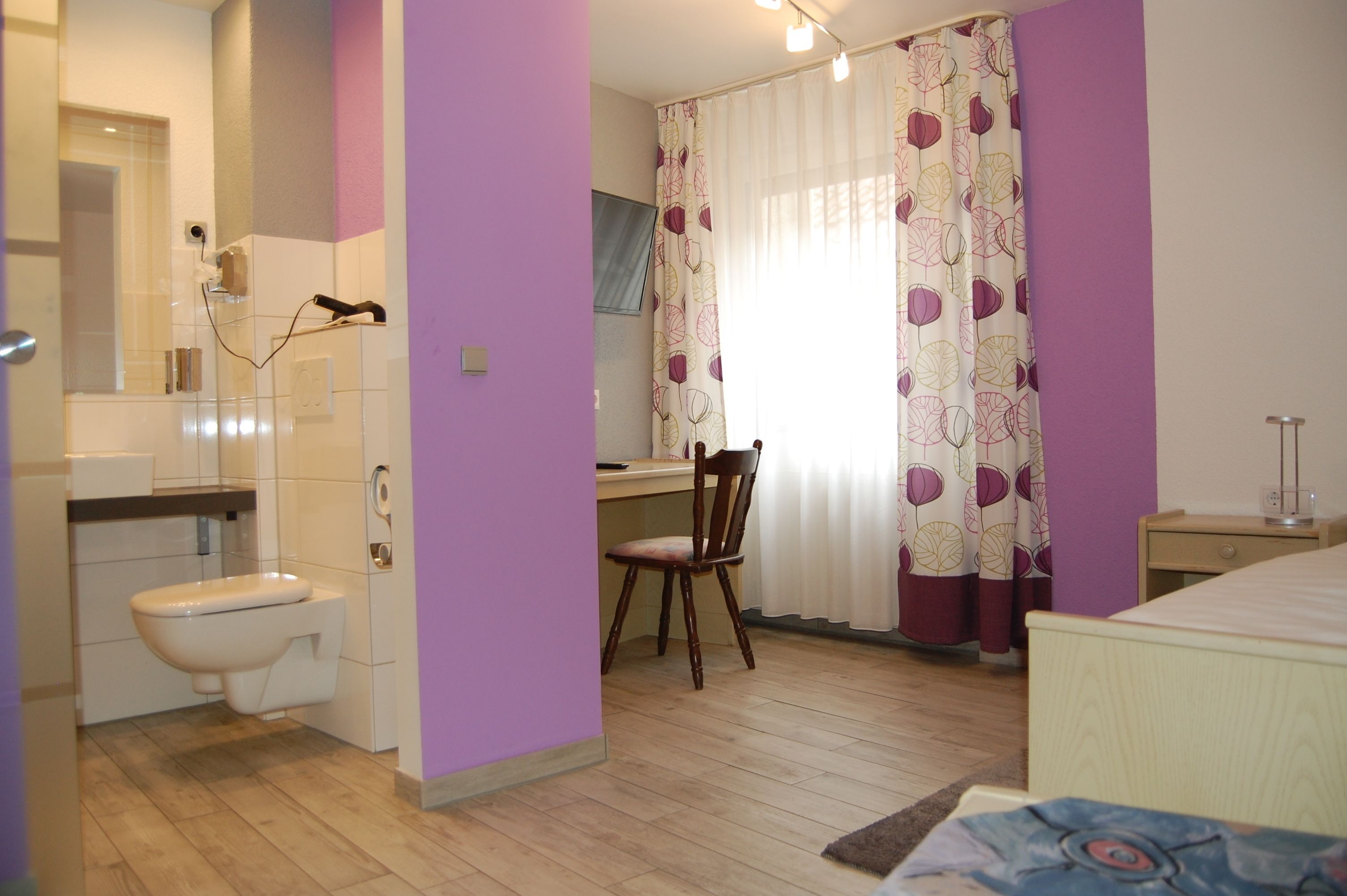 A bathroom with a toilet, sink, and shower. There is a chair and a desk with a chair in the room.