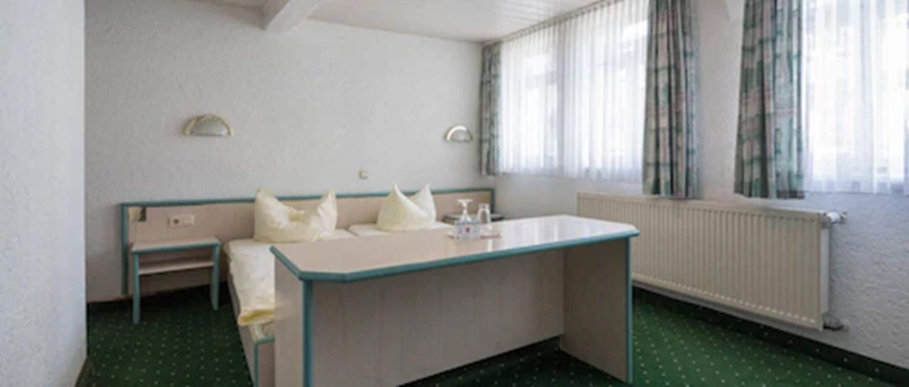Una habitación pequeña y sencilla con una cama individual, una mesita y un ventanal con cortinas.