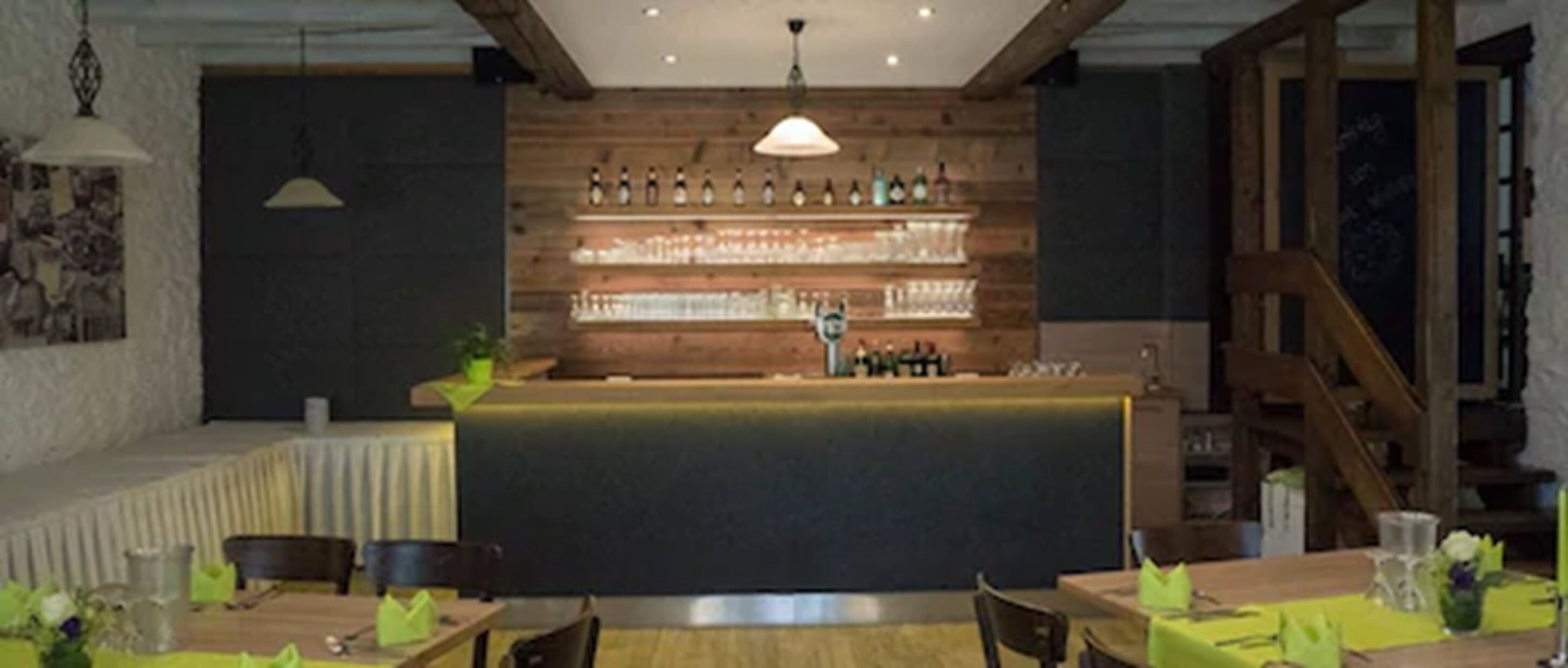 Un restaurant amb barra de fusta, taules amb mantells verds i lluminàries penjants.