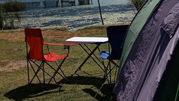 Un campement comprenant une tente, deux chaises et une table, installé près d’un lac où l’on peut voir des bateaux, avec une montagne boisée en arrière-plan.
