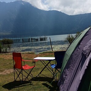 A camping setup with a tent, two chairs, and a table near a lake with boats and a forested mountain in the background. - Bedugul Camping (Baturiti)