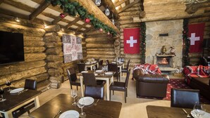 A cozy log cabin interior with wooden walls, stone fireplace, and festive decorations. Tables are set for dining with wine glasses and cutlery. - Monastic-SPA (Oriv)