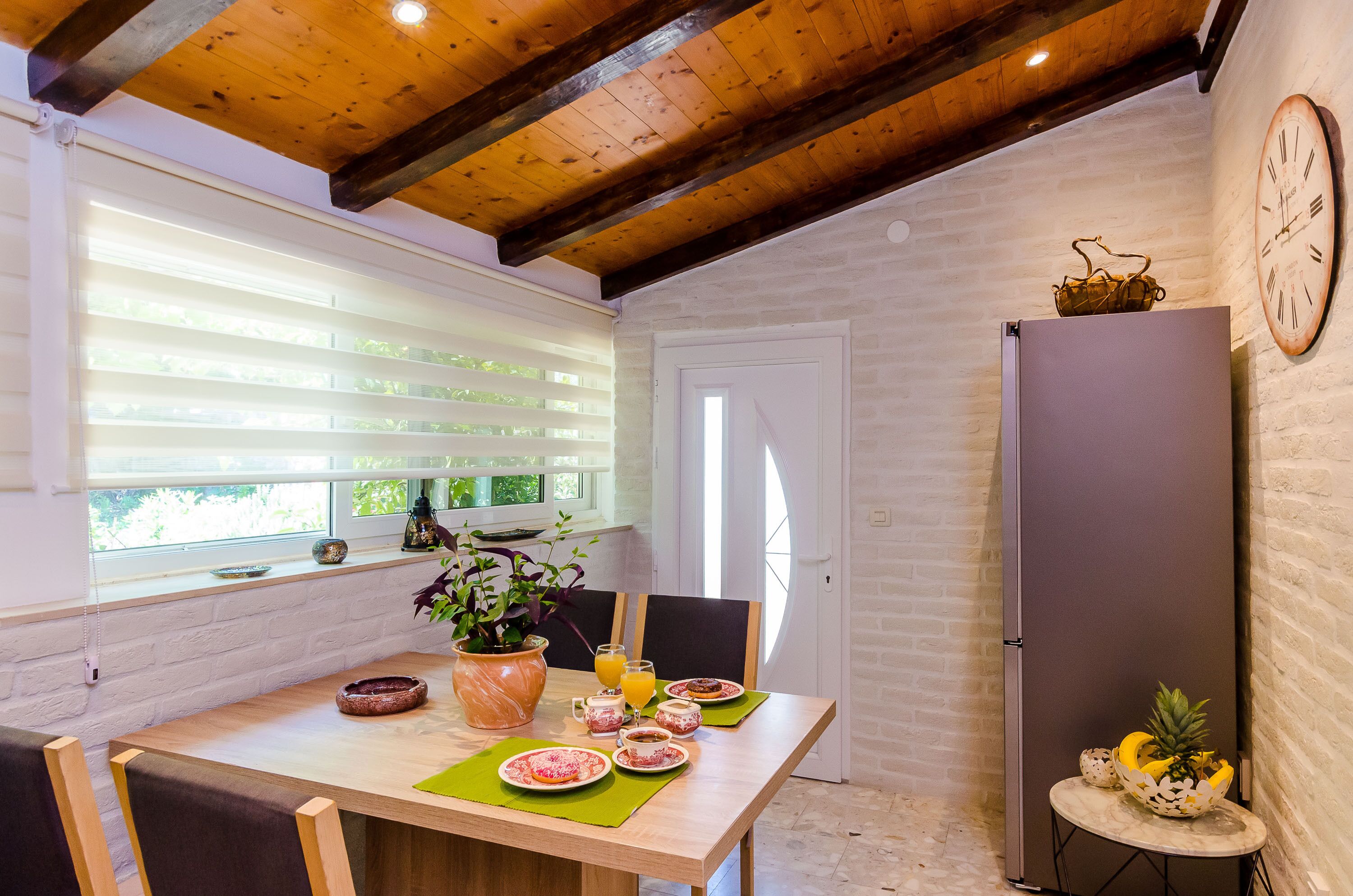 A dining area with a wooden table, chairs, a potted plant, and a clock on the wall.