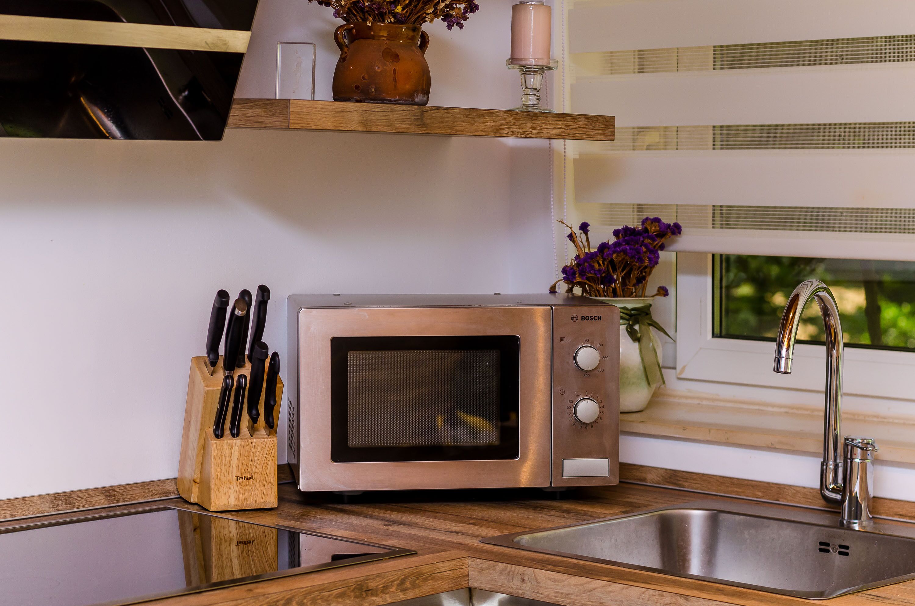 A kitchen with a stainless steel microwave, a knife block, a vase with flowers, and a window with a view of greenery.