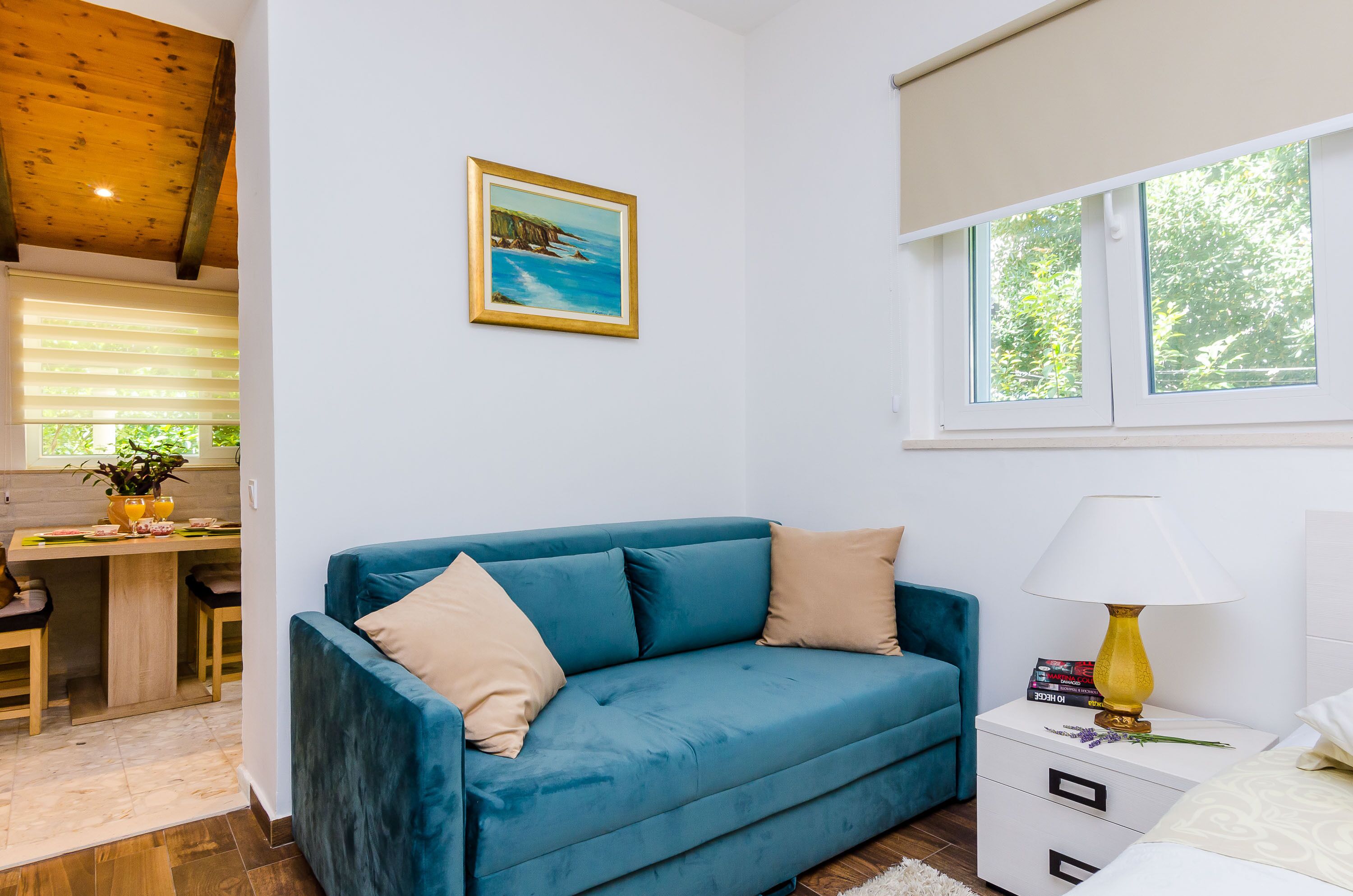 Een moderne woonkamer met een blauwe bank, een klein wit nachtkastje met een lamp, en een schilderij van een kustlandschap.